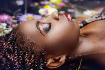 Wall Mural - Glamorous African woman with bright makeup on the eyes closed lying in the water with flowers close-up view of the profile