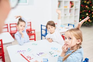 three little children with raised hands sitting by table and looking to their teacher at lesson in k