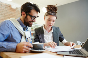 Wall Mural - Young confident manager and her colleague reading business contract by table during coffee-break in cafe