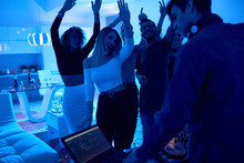 Group Of Modern Young People Dancing Listening To DJ Playing Music At Private House Party, Lit By Blue Light