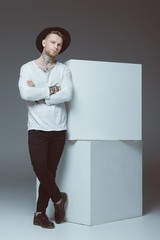 stylish young man with tattoos standing with crossed arms and looking at camera on grey