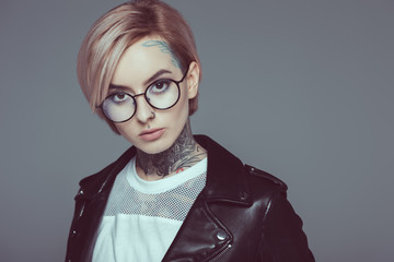 tattooed girl with pink hair in eyeglasses and black leather jacket, isolated on grey