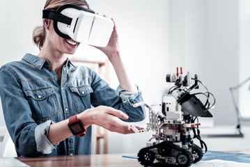 Under reality. Young woman touching virtual mask and expressing positivity while looking at robot