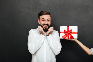 Sticker - Portrait of excited brunette man rejoicing to get white gift box with red bow from female hand, over dark gray wall