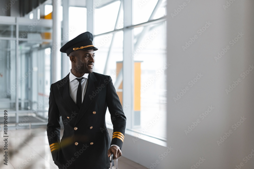 handsome young pilot with luggage at airport looking away - obrazy, fototapety, plakaty 