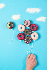 Wall Mural - Colored mini donuts on blue background, creative food idea, hand holding donuts in a shape of balloons in the sky with clouds made of coconut, top view