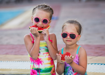 Two kids in swimming suits sitting in swimming pool and eating red watermelon