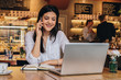 © foxyburrow - Young businesswoman dressed in white shirt is sitting in cafe at wooden table in front of laptop and talking on cell phone. Telephone conversations. Hipster girl is talking on phone with her friend.