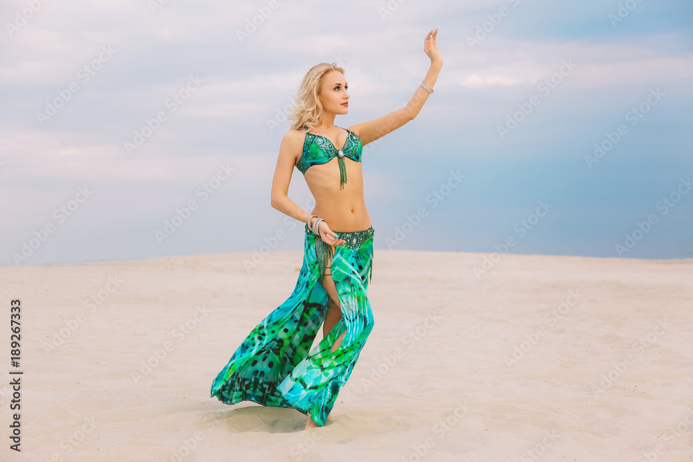 Foto de Stock A beautiful blond girl in a golden with green dress with  wings, suit is dancing an oriental, East dance in the desert on the sand.  Beautiful exotic woman dancer