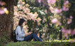 © NAMPIX - Beautiful young woman sitting under big tree on sunny spring day in park during cherry blossom season, reading a book
