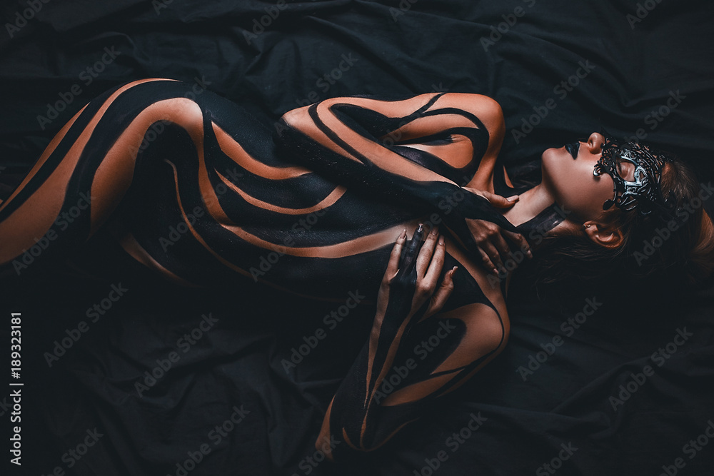 Beautiful girl with painted body and black mask in her face lies on bed. Body art. - obrazy, fototapety, plakaty 