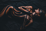 Beautiful girl with painted body and black mask in her face lies on bed. Body art.