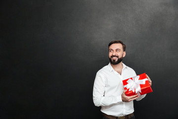 Poster - Picture of smiling bearded man holding red gift box with white ribbon and looking aside, over dark gray wall