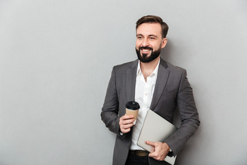 Poster - Portrait of cheerful male office worker posing on camera holding takeaway coffee and silver laptop, isolated over gray wall