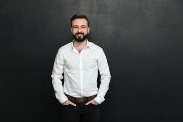 Poster - Portrait of young man in white shirt posing on camera with broad smile, and hands in pockets over dark gray background