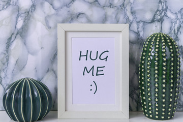 Ceramic cactus and white frame with the inscription hug me on a white desk on the background of a marble wall