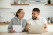 © fizkes - Young smiling happy couple talking at home in the kitchen with laptop papers, cheerful man and woman holding documents excited by planning new purchase, having fun enjoying positive news in letter