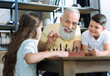 © zinkevych - Family leisure time. Selective focus on a funny retired man and his grandson smiling cheerfully and looking at a cute little girl while all playing chess together.