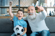 © zinkevych - Hurray. Extremely happy senior gentleman and his grandson sitting on a sofa and lifting their arms with excitement while watching a football match together.