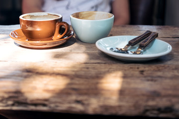 Two cups of hot coffee and a plate with spoon and fork on vintage wooden table with people in background