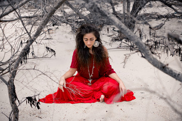 Wall Mural - Beautiful young woman in red dress sitting on sand on the beach. Witchcraft conceptual image