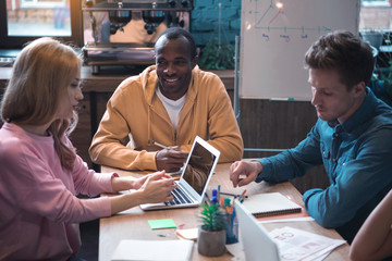 Poster - Portrait of cheerful man speaking with side view calm woman while locating at desk. She typing in laptop. Serious comrade situating opposite her. Work concept