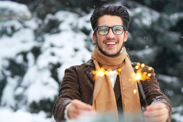 Outdoor close up portrait of young beautiful men with nice hair. Smiling young man having fun with sparklers. Man enjoy a winter. Dressed in a coat, sweater and scarf .winter concept. Snowfall.