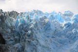 Fototapeta  - ostre krawędzie czoła lodowca w kolorze biało niebieskim w zbliżeniu z zachmurzonym niebem w tle