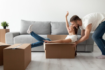 Happy couple having fun laughing moving into new home, young excited woman riding sitting in cardboard box while man pushing it, cheerful roommates playing while packing unpacking belongings together