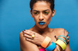 Colorful photo of tense or disappointed mixed-race woman with trendy makeup and accessories posing with crossed hands on chest, over blue wall