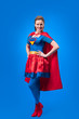 © LIGHTFIELD STUDIOS - beautiful smiling woman in superhero costume standing akimbo isolated on blue