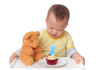 Wall Mural - Cute little Asian 2  years old toddler baby boy child sitting in high chair celebrating birthday and blowing candle on red velvet cupcake with teddy bear, isolated on white with clipping path