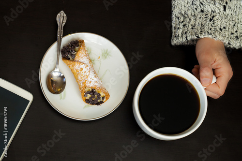 Fotografie, Obraz Caucasian woman hand holds coffee cup with smartphone and italian cannoli on pla