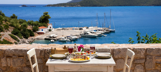 A table served for two with chicken souvlaki and french fries, greek salad, snacks and drinks on the summer terrace of the hotel room by the seascape, beautiful summer greek holidays concept.