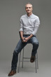 Man in a shirt and jeans is sitting on a chair.
Gray background.