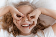 © LIGHTFIELD STUDIOS - top view of smiling adorable child lying on bed and showing eyeglasses gesture