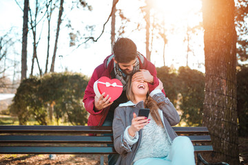 Wall Mural - Handsome man surprising his girlfriend with a gift. Young man with giftbox closing his girlfriend eyes to make a surprise for her.