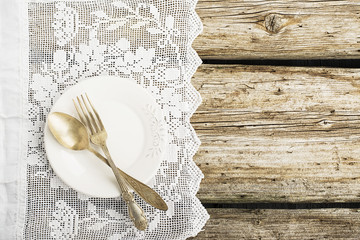 White ceramic plates on a simple wooden background with a white vintage lacy tablecloth with silverware table instruments with a pink paper heart. Top view. Copy space,