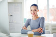 © pressmaster - Happy girl with toothy smile sitting by desk in office in front of computer monitor and looking at camera