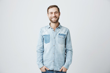 Friendly looking positive Caucasian stylish man with stylish haircut and bristle smiling broadly during nice conversation with good friend, standing isolated against grey studio wall.