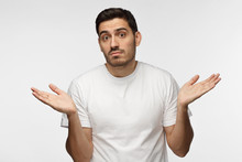 I Don't Know. Young Man Isolated On Grey Background Being At A Loss, Showing Helpless Gesture With Arm And Hands, Mouth Curved As If He Does Not Know What To Do