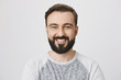 Cute bearded guy with shiny and cheerful smile looking at camera while standing against gray background. Adult european model promotes new item that will go viral on market.