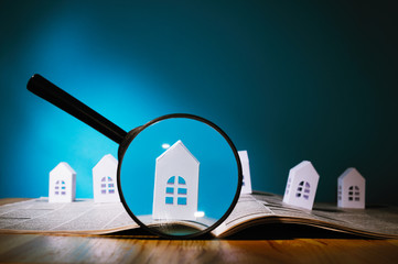 Magnifying glass in front of an open newspaper with paper houses. Concept of rent, search, purchase real estate.