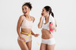 © Yakobchuk Olena - Joyful ladies keeping bodies in shape. African woman using measure tape, another one holding sports bottle and towel on her neck. Isolated on background