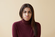 Portrait of upset young mixed race woman in maroon turtleneck pouting, feeling disappointed and unhappy after quarrel with her boyfriend, posing in studio with copyspace wall for your advertisement