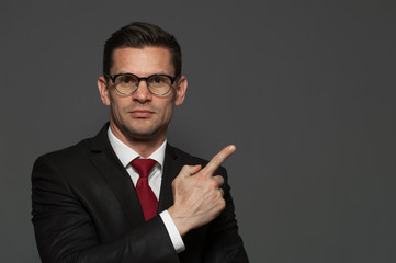 Wall Mural - Portrait of successful young positive businessman pointing his finger at the space showing something useful on gray background