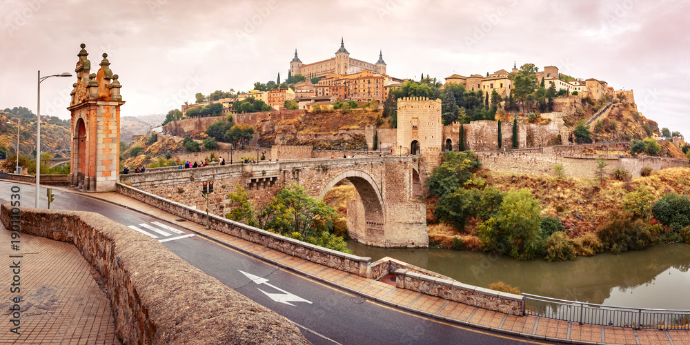 Panorama of Old city of Toledo with Alcantara Bridge across the river Tajo and Alcazar in the overcast evening, Castilla La Mancha, Spain - obrazy, fototapety, plakaty 