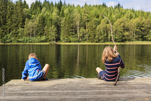 Fotografie, Obraz Two children using a homemade fishing rod fishing from a jetty by a lake set in an idyllic Swedish summer forest landscape