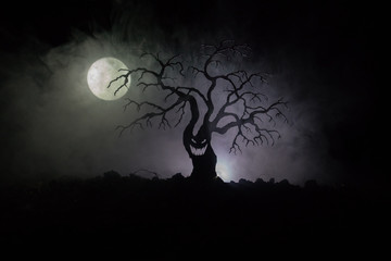 Wall Mural - Silhouette of scary Halloween tree with horror face on dark foggy toned background with moon on back side. Scary horror tree with zombie and demon faces.