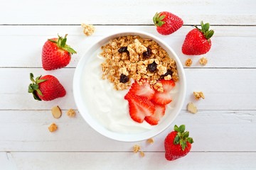 Wall Mural - bowl of yogurt with strawberries and granola, over a white wood background. scattered berries.